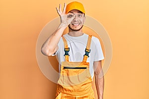 Hispanic young man wearing handyman uniform doing ok gesture with hand smiling, eye looking through fingers with happy face