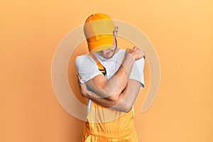 Hispanic young man wearing handyman uniform hugging oneself happy and positive, smiling confident