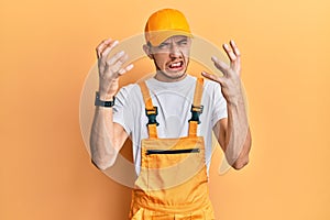 Hispanic young man wearing handyman uniform shouting frustrated with rage, hands trying to strangle, yelling mad