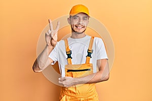 Hispanic young man wearing handyman uniform smiling looking to the camera showing fingers doing victory sign