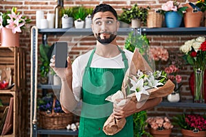 Hispanic young man working at florist shop showing smartphone screen smiling looking to the side and staring away thinking