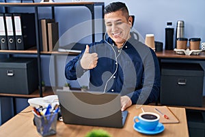 Hispanic young man working at the office with laptop smiling happy and positive, thumb up doing excellent and approval sign