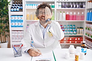 Hispanic young man working at pharmacy drugstore covering one eye with hand, confident smile on face and surprise emotion