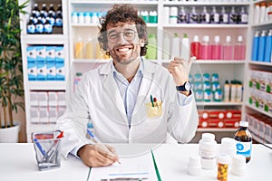 Hispanic young man working at pharmacy drugstore pointing to the back behind with hand and thumbs up, smiling confident