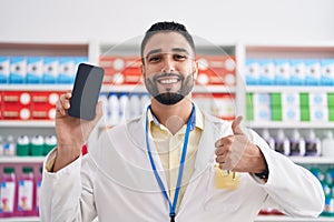 Hispanic young man working at pharmacy drugstore showing smartphone screen smiling happy and positive, thumb up doing excellent