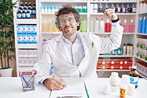 Hispanic young man working at pharmacy drugstore strong person showing arm muscle, confident and proud of power