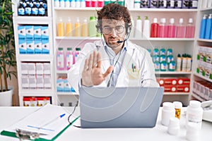 Hispanic young man working at pharmacy drugstore working with laptop with open hand doing stop sign with serious and confident