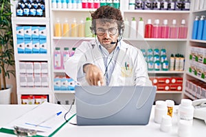 Hispanic young man working at pharmacy drugstore working with laptop pointing with finger to the camera and to you, confident