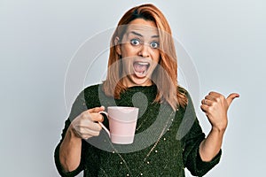 Hispanic young woman drinking a cup coffee pointing thumb up to the side smiling happy with open mouth