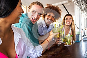 Hispanic young woman and multiracial friends toasting with mojito cocktails at a beach bar.