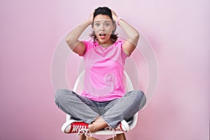 Hispanic young woman sitting on chair over pink background crazy and scared with hands on head, afraid and surprised of shock with