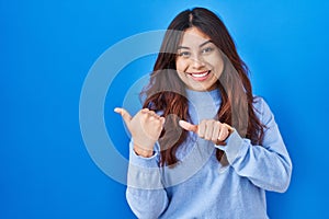 Hispanic young woman standing over blue background pointing to the back behind with hand and thumbs up, smiling confident