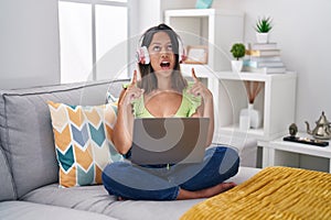 Hispanic young woman using laptop at home amazed and surprised looking up and pointing with fingers and raised arms