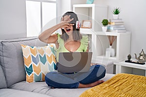 Hispanic young woman using laptop at home peeking in shock covering face and eyes with hand, looking through fingers with