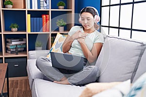 Hispanic young woman using laptop at home wearing headphones smiling with hands on chest with closed eyes and grateful gesture on