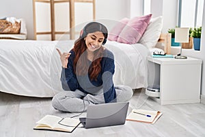 Hispanic young woman using laptop sitting on the floor at the bedroom smiling happy pointing with hand and finger to the side