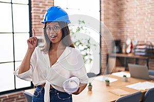 Hispanic young woman wearing architect hardhat at office surprised with an idea or question pointing finger with happy face,