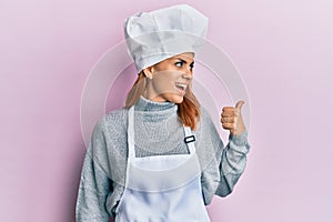 Hispanic young woman wearing professional cook uniform and hat pointing thumb up to the side smiling happy with open mouth