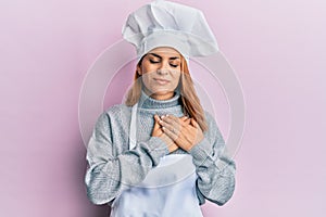 Hispanic young woman wearing professional cook uniform and hat smiling with hands on chest, eyes closed with grateful gesture on