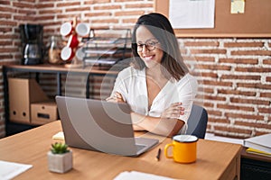 Hispanic young woman working at the office wearing glasses happy face smiling with crossed arms looking at the camera