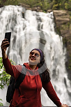 Hispoanic woman taking selfie with mobile at lookout of waterfall. Travelers concept