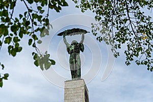 The historic Liberty Statue upon Gellert Hill in front of a cloudy sky and green leaves in Budapest, Hungary