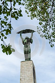 The historic Liberty Statue upon Gellert Hill in front of a cloudy sky and green leaves in Budapest, Hungary