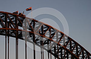 Historic steel truss arch bridge with crane, Australian and New South Wales flags on top. Iconic Sydney Harbor Bridge at dusk.