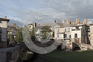 a historic town with stone buildings, an arched bridge, and distant castles under a partly cloudy sky