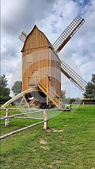 Historic windmill made of light brown wood . Wooden building with barrier band , green grass and very white cloudy sky . Ancient