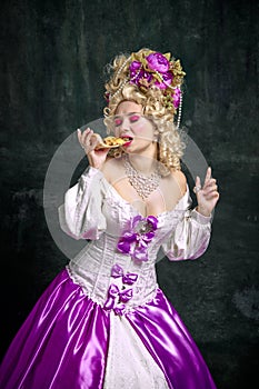 Historical portrait of young attractive woman in dress with corset eating delicious piece of pizza against vintage dark