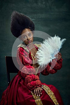 Historical reenactment. Medieval royal person, young african woman, queen in stylish dress with fan sitting against