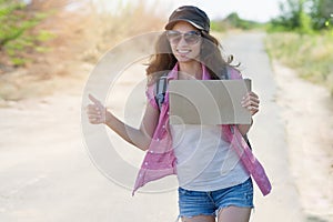 Hitchhiking woman travel. Beautiful young female hitchhiker in a cap and sunglasses smiles and catches a passing car by