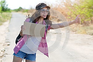 Hitchhiking woman travel. Beautiful young female hitchhiker in a cap and sunglasses smiling while standing by the road during