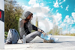 Hitchhiking. A young woman in cap and sunglasses sitting along the road, holding a paper map and looking at driving car