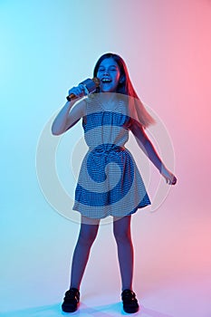 Hobby. Little talented girl, child singing in microphone over gradient blue pink studio background in neon light