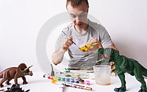 Hobby: a man paints dinosaur toy figures with a brush and paints on a white table.