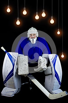 Hockey goalie in complete hockey outfit sitting on office chair. Above him are lamps with a light bulb on.