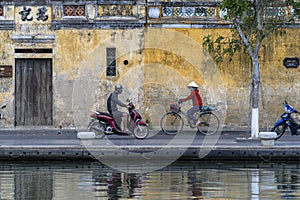 Asian woman rides a bicycle and a man rides a motorbike the road near the river in the old city against the background of the old