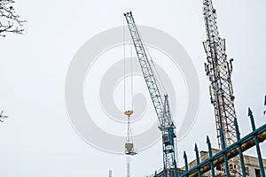 The Hoisting Tower Crane Moves the Concrete Mixer to The Workplace at The Construction Site. A Cell Phone Mast Is Visible Next to