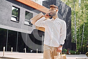 Holding bags and talking by phone. Stylish young man have shopping day. Outdoors with bags