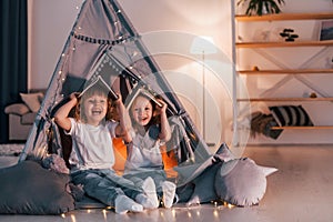 Holding books on head. Playful mood. Two little girls is in the tent in domestic room together