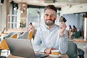 Holding credit cards. Young man is sitting in the cafe and working by using laptop