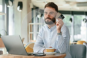 Holding credit cards. Young man is sitting in the cafe and working by using laptop