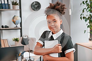 Holding notepad and smiling. Cute little girl in school uniform is standing indoors in domestic room