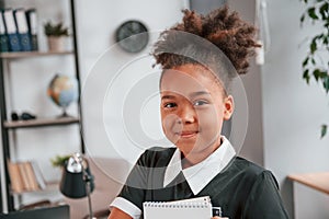 Holding notepad and smiling. Cute little girl in school uniform is standing indoors in domestic room