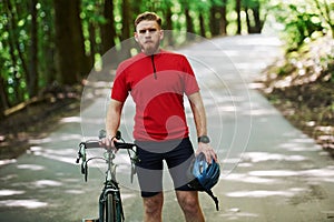 Holding safety helmet in hand. Cyclist on a bike is on the asphalt road in the forest at sunny day