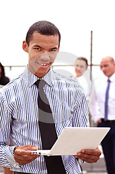 Holding a tablet computer, a young handsome black businessman is standing outside of a business building, confidently