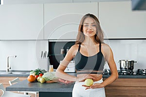 Holds apple against belly. Young european woman is indoors at kitchen indoors with healthy food