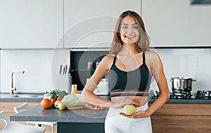 Holds apple against belly. Young european woman is indoors at kitchen indoors with healthy food